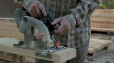 Yuvarlak testereli yaşlı bir adamın ellerine yakın çekim. Adam açık hava atölyesinde odun kesmek için ustaca dairesel testere kullanıyor..