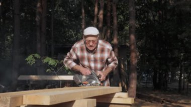 Yaşlı bir zanaatkar, ormanın huzurlu güzelliğinin ortasında dairesel bir testereyle tahta kesmeye odaklanıyor. Güneş ışığı ağaçların arasından süzülür, atmosferi güçlendirir..