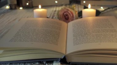 Davetkar bir okuma alanı, parlak mumlar ve narin bir gülle çevrili açık bir kitap içerir. Rahatlama ve yansıma için mükemmel sıcak bir atmosfer yaratır..