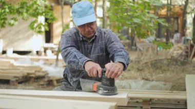 Yetenekli bir işçi, yeşillik ve doğal ışıkla çevrili bir açık hava projesi için ahşap kalasları şekillendiren bir zımpara makinesini dikkatlice kullanıyor. Atmosfer sakin ve odaklı..