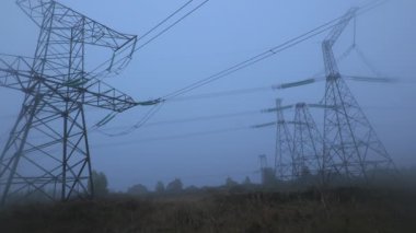 Kırsal kesimde sabahın erken saatlerinde, yoğun sis yüzünden kısmen gizlenen elektrik hatlarının atmosferik ve sakin bir manzara yarattığını gösteriyor. Sahne, doğanın sessizliğini yakalar..