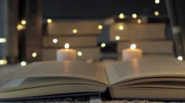 Açık kitap, yanan mumlar ve arka planda peri ışıklarıyla odak değiştiriyor. Arka planda bir yığın kitap bulanıklaştırılmış