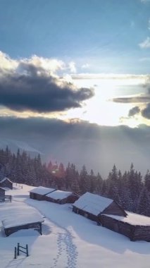 Sakin bir kış manzarası karlı bir alana yerleşmiş sade ahşap kulübeler sergiliyor, güneş ışığı bulutların arasından gün batımında doğuyor, durgun manzarayı aydınlatıyor..