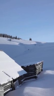 İki kırsal kulübe, gün ortasında açık mavi bir gökyüzü altında, kayan tepeler ve ebedi yeşil ağaçlardan oluşan huzurlu bir kış manzarasıyla çevrili kalın bir kar tabakasının altında yer alır. Hava görüntüsü, dikey görüntüler