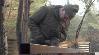 Bir zanaatkar ağaçlık alanda öğütücüyle metal kesmeye odaklanır. Maddeyi şekillendirirken kıvılcımlar uçuşuyor, doğadaki sanatına bağlılığını gösteriyor..