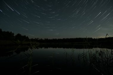 Gecenin karanlığında, yıldız izleri sakin bir gölün üzerinde büyüleyici bir görüntü yaratır. Durgun su, ışığı ve etrafındaki ağaçları yansıtır ve sakin atmosferi güçlendirir..