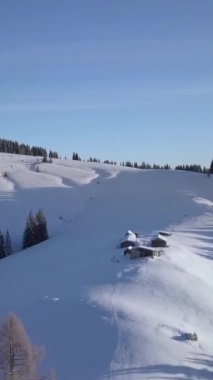 Yuvarlanan karla kaplı tepelerin olduğu sakin bir kış manzarasının havadan görüntüsü. Kar ve ağaçların arasına yerleştirilmiş küçük bir kulübe bu uzak yerin sakinliğini gösteriyor. Dikey görüntüler