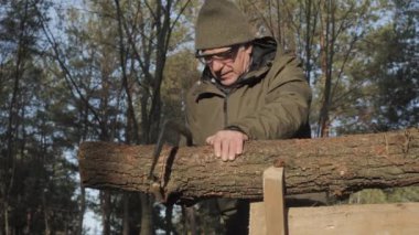Sıcak giysili yaşlı bir adam, uzun ağaçlar ve doğal ışıklarla çevrili huzurlu bir orman ortamında, bir kütükle titizlikle çalışıyor..
