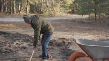 Yeşil ceketli ve şapkalı bir adam inşaat alanında toprağı el arabasına taşımak için kürek kullanıyor.