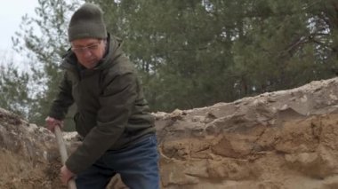 Bir adam kış boyunca ormanlık bir alanı kazıyor, kalın bir ceket ve şapka giyiyor. İnşaat veya peyzaj projesi için zemin hazırlamak için kürek kullanıyor..