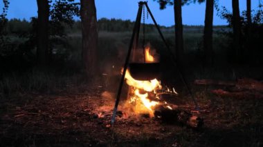 Büyük bir tencere alacakaranlıkta sakin bir orman ortamında çatırdayan bir kamp ateşinin üzerinde asılı duruyor. Yemek pişerken alevler kararan gökyüzüne çarpıyor..