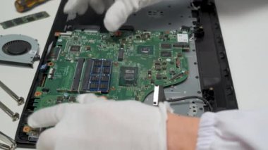 Beyaz eldiven giyen bir teknisyen, bir atölyede hassasiyet ve temizlik sağlamak için, onarım işlemi sırasında bir dizüstü bilgisayarın ana kartını dikkatle tutuyor..