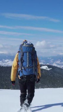 Sarı ceketli ve mavi sırt çantalı bir yürüyüşçü, parlak mavi bir gökyüzünün altında görkemli dağlarla çevrili derin karda yürüyor. Manzara nefes kesici bir şekilde temiz. Dikey görüntüler