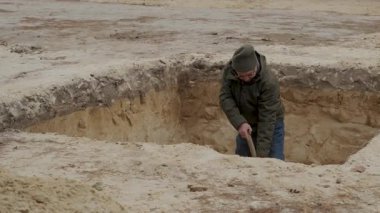 Şapka ve ceket giyen bir adam kumlu zeminde derin bir çukur kazıyor. Bu kazı projesinde özenle çalışırken toprak yığınları bir kenara atılıyor..