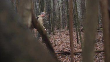 Bir insan uzun ağaçlar ve yapraklarla dolu sık bir ormanda bir elinde baltayla dolaşır ve ağır bir sırt çantası taşır. Açık hava macerası izlenimi uyandırır..