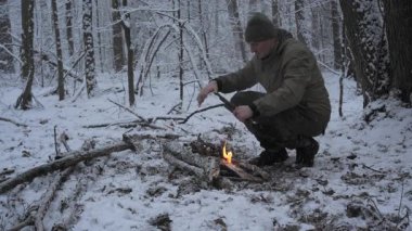 Bir hayatta kalma meraklısı, karla kaplı bir ormanda ateş yakmak için çubukları dikkatlice yerleştirir. Sakin kış ortamı kar tanelerinin arasında sakin bir atmosfer yaratıyor..