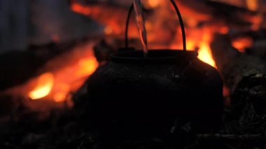 Su, kamp ateşinin üzerinde duran siyah bir çaydanlığa dökülüyor. Alevlerin sıcaklığı çevreyi aydınlatarak doğada rahat bir atmosfer yaratıyor..