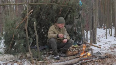 Bir adam kış ormanında kamp ateşinin yanında oturuyor. O, çam dallarından bir sığınak inşa etti ve sıcak açık hava giysileri giydi. Sahne hayatta kalmayı, çalı ustalığı becerilerini yansıtıyor..