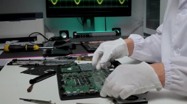 Beyaz eldiven ve laboratuvar önlüğü giyen bir teknisyen çalışma masasındaki dizüstü bilgisayarı dikkatlice onarıyor. Arka planda bir dalga biçimini gösteren elektronik araçlar ve bir monitör var.