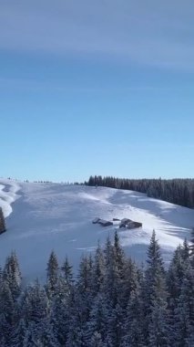 Hava manzarası, karla kaplı tepeler ve berrak mavi bir gökyüzü altında sık ve yeşil ağaçların arasına yerleşmiş izole edilmiş kulübelerin yer aldığı sakin bir kış manzarası yakalar. Dikey görüntüler