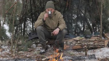 Bir adam kış ormanında kamp ateşinin yanında oturuyor. O, çam dallarından bir sığınak inşa etti ve sıcak açık hava giysileri giydi. Sahne hayatta kalmayı, çalı ustalığı becerilerini yansıtıyor..