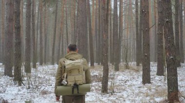 Sırt çantalı bir gezgin karlı bir çam ormanında yürüyor. Açık havada hayatta kalma kıyafeti giymiş, hasırla sarılmış, maceraya hazır..