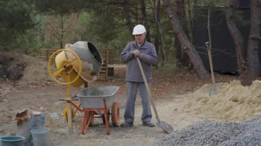 Miğfer takan yaşlı bir inşaat işçisi elinde kürekle beton karıştırıcının yanındaki şantiyede dinleniyor.