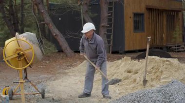 Yaşlı inşaat işçisi çimento karıştırıyor, miğferli yaşlı adam inşaat alanında beton karıştırıcının yanında kürekle çalışıyor..