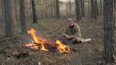 Ormanda kamp ateşinin yanında oturan adam Bushcraft Survival 'da çalışıyor, çam ormanında tek başına ısınan kampçı, barışçıl orman yaşamının tadını çıkarıyor.