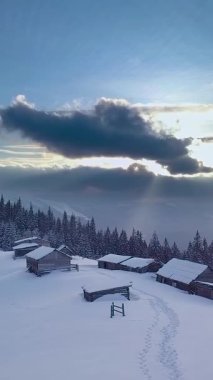 Kar, sakin bir kış manzarasında kırsal kabinleri örtüyor. Yukarıda kara bulutlar beliriyor, yumuşak güneş ışığıyla zıt düşüyor ve sakin bir atmosfer yaratıyor. Dikey görüntüler