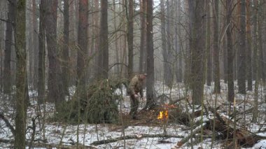 Karlı bir ormanda kamp ateşi olan bir adam kar tozlu bir ormanda çalı çırpı becerileri geliştiriyor, barınak inşa ediyor ve bir yangına bakıyor.