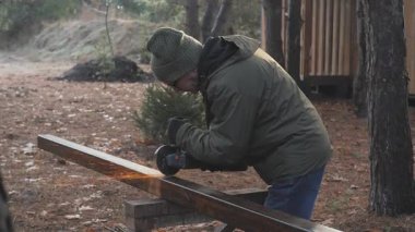 Bir insan ağaçlar ve sonbahar yapraklarıyla çevrili sakin bir orman ortamında ahşap bir kalas zımparalıyor. Sabahın erken saatleri ahşap işçiliği için huzurlu bir atmosfer yaratır..