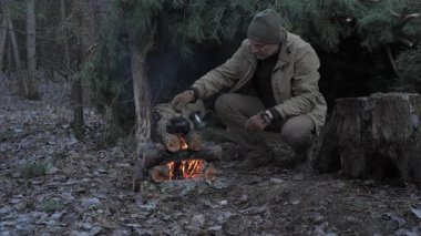Bir adam ormanda ateşin üzerinde yemek pişiriyor, çam dallarının yanına çömeliyor, ormandaki yanan odunların üzerine çaydanlık koyuyor..