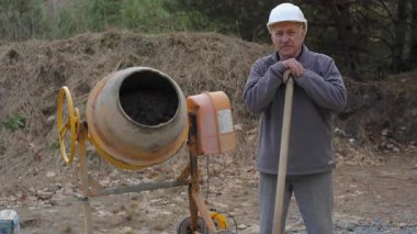 Kıdemli İnşaat İşçisi Beton Karıştırıcı 'nın yanında duruyor. Kasketli ve iş kıyafetli yaşlı bir adam inşaat sahasında beton karıştırıcının yanındaki küreğin üzerinde dinleniyor..