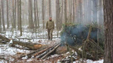 Karlı Orman 'da Smoky Bushcraft Barınağı' na yaklaşan bir adam, açık hava kıyafetleri giymiş bir adam sisli, karla kaplı bir ormandaki bir orman sığınağına doğru yürüyor..