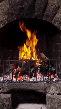 Alevli dikey taş şömine, canlı alevi olan taştan bir şömineye yakın çekim ve metal ızgaranın arkasında parlayan közler. Dikey görüntüler