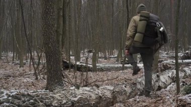 Bir adam, bir kış günü, bir sırt çantasıyla düşmüş bir kütük boyunca, karlı bir ormanda gezinir. Manzarada çıplak ağaçlar ve kar parçaları yer alıyor..