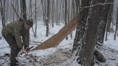 Bir insan, karlı bir ormanlık alanda branda ve dallar kullanarak geçici bir barınak inşa eder. Çevre soğuk ve sakin, kar taneleri yavaşça düşüyor..