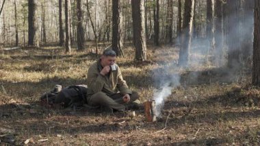Ormandaki Kamp Ateşi 'nin İçen Adam, orman zemininde oturmuş sigara içen küçük bir kamp ateşinin yanında fincandan içki içen adam..