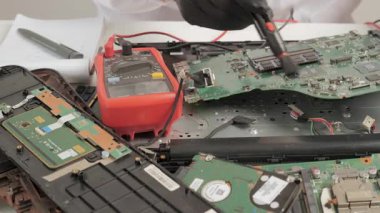 Yakınında çoklu metre olan teknisyen temizlik ana kartı, parçalanmış dizüstü bilgisayarı ve arka planında not defteri olan elektronik devre bakımı.