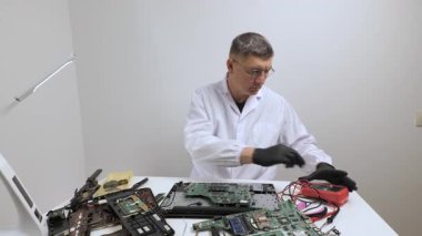 Laboratuvar önlüklü bir elektronik teknisyen, çalışma masasında çoklu metre kullanarak bir laptop ana kartını test ediyor..
