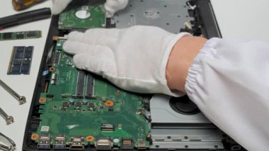Beyaz eldiven giyen teknisyen, beyaz bir masadaki çalışma alanını çevreleyen çeşitli araçlarla bir dizüstü bilgisayar ana kartı bileşenini dikkatlice yerleştirir veya ayarlar