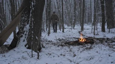 Karla kaplı uzak bir ormanda, bir adam, uzun ağaçlar ve kış manzarasıyla çevrili küçük bir yangını körüklemek için odun toplar ve yalnızlık hissi uyandırır..