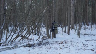 Kereste taşıyan Ormanda Kış Çalılığı, haki rengi ceket ve yeşil pantolon giyen bir adam karlı bir orman sahnesinde omzunda büyük bir kütük taşıyor.