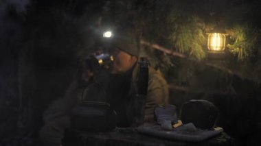 Gece Bushcraft Kamp Ateşi Çam Barınağı Altında, elinde lamba olan bir adam gece fenerle yanan bir çalılık barınağında ateşin yanındaki metal bardaktan su içiyor.