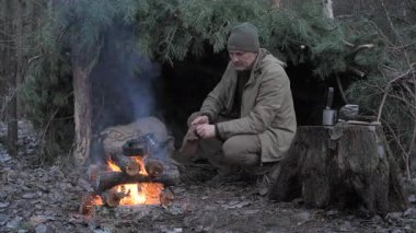 Soğuk ormanda çam ağacının yanında çay hazırlayan bir adam çöldeki kırsal barınağında dinlenirken bir kampçı, ateşte çaydanlığa bakar.