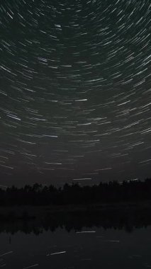 Gölün üzerindeki gece gökyüzünde yıldız izleri zaman aşımına uğradı. Sudaki yıldızların yansıması. Destansı video, dikey görüntü