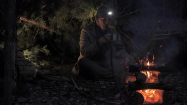 Gece Bushcraft Kamp Ateşi Çam Barınağı Altında, elinde lamba olan bir adam gece fenerle yanan bir çalılık barınağında ateşin yanındaki metal bardaktan su içiyor.