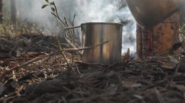 Orman kamp alanındaki metal bardağa sıcak su döken buharlı içecek, kırsal alanda hayatta kalma mücadelesi sırasında orman zemininde teneke bardağa akıyor.