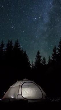 Yıldızlı gökyüzünün altında gece vakti, uzun çam ağaçlarıyla çevrili, yıldızlarla ve çam ağaçlarıyla dolu bir gökyüzü ve dikey görüntülerle dolu, karanlık bir ormanda parlayan kamp çadırı.